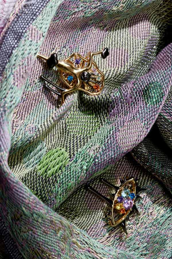 Close-up view of two ornate, gold statement rings set with colourful gemstones, pinned onto the folds of a textured green and purple woven fabric.