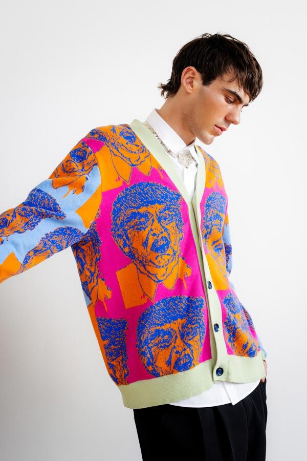 Male model looking down while wearing a vibrant pink, orange, and blue cardigan featuring a stylized graphic portrait print, styled over a white shirt and black trousers against a plain white background.