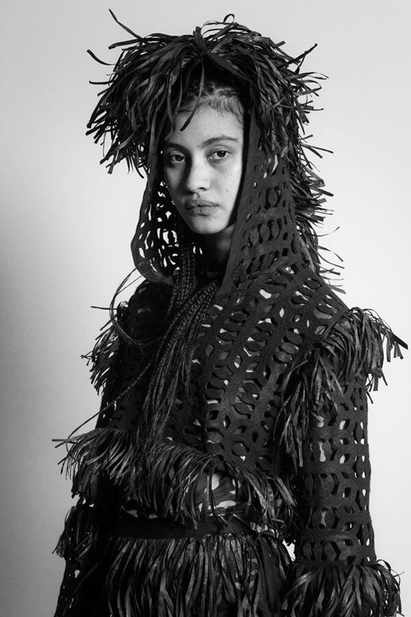 Black and white studio portrait of a model wearing a textured, open-knit hooded jacket featuring heavy fringe detailing along the sleeves, waist, and around the head.