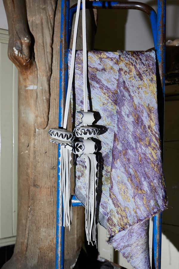 A textured, shimmering purple and gold metallic fabric draped over a blue metal frame next to a white leather tasselled hanging accessory.