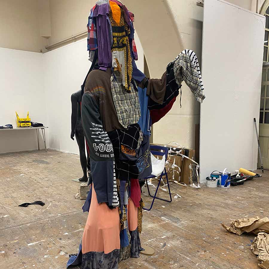 A large, abstract sculpture composed entirely of clothing. The sculpture resembles a human figure with multiple limbs and a head covered in layers of fabric