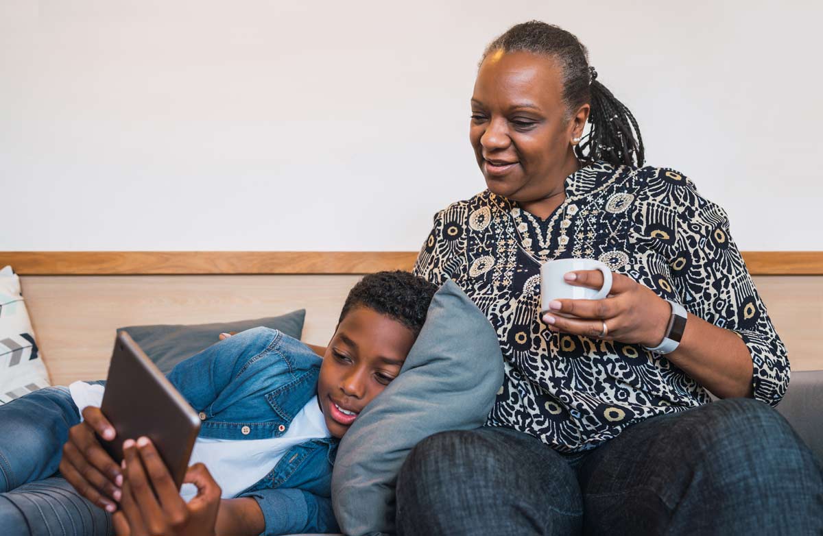 An older black woman with a touch of grey hair at her temples and braids sits on a sofa with a young black boy looking at a digital tablet. She's drinking a cup of tea and they look relaxed and happy.