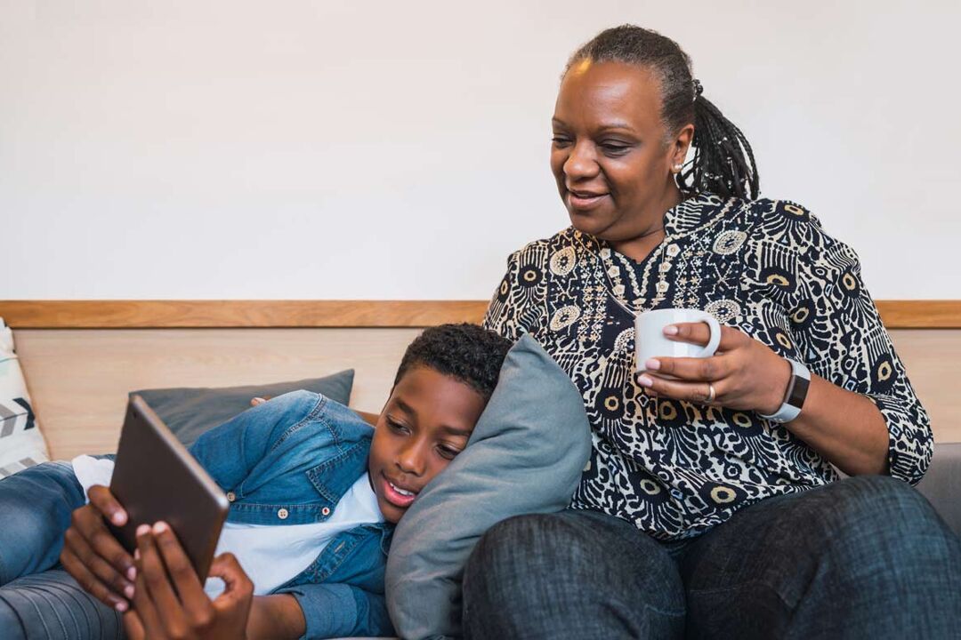 An older black woman with a touch of grey hair at her temples and braids sits on a sofa with a young black boy looking at a digital tablet. She's drinking a cup of tea and they look relaxed and happy.