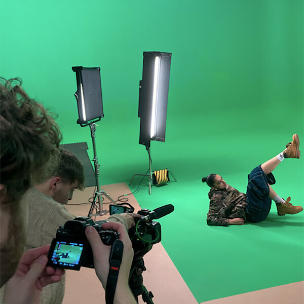 Student sat on floor with legs raised, using a green screen, with two students taking pictures