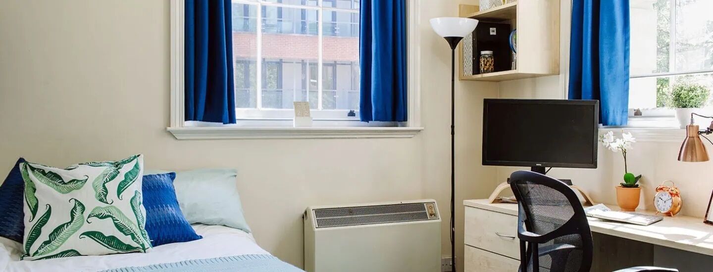 A tidy bedroom with blue curtains and cushions. A comfy looking bed sits on one side of the room with a neat desk for studying on the other. 