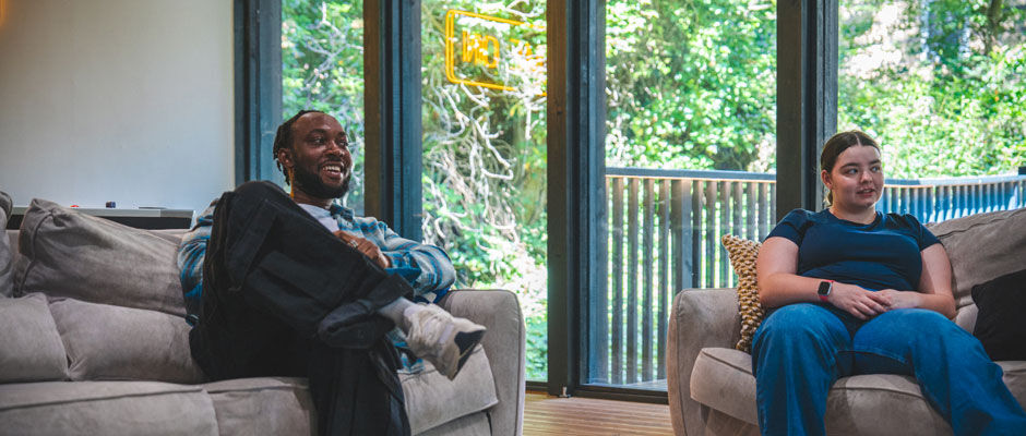 Two students relaxing in the common room of their accommodation. They sit on two sofas, laughing and talking.