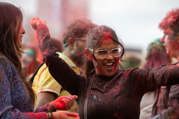 A student dances outside at the Holi festival. She is covered in red paint an dis surrounded by other students covered in paint of different colours.