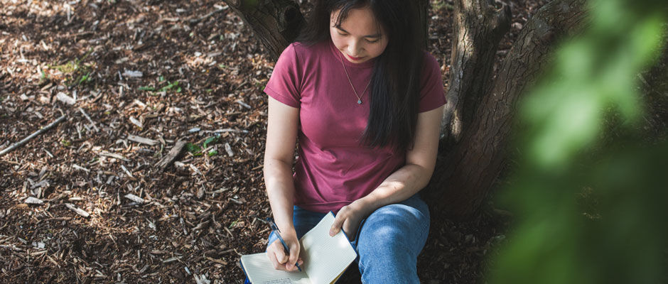 A student sitting outside by a tree writing in a notebook