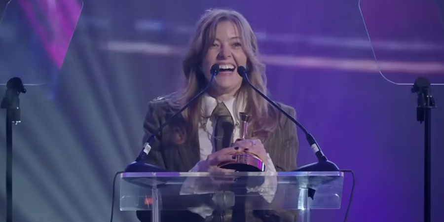 A young woman standing on a clear podium onstage, smiling widely while holding an award. She is framed by two microphones and lit by purple and blue stage lights in a celebratory atmosphere.
