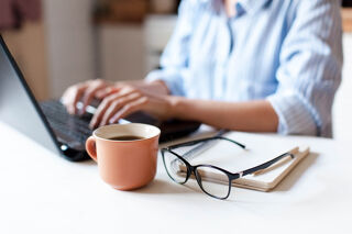 A person typing on a laptop with some glasses and a cup of coffee near by