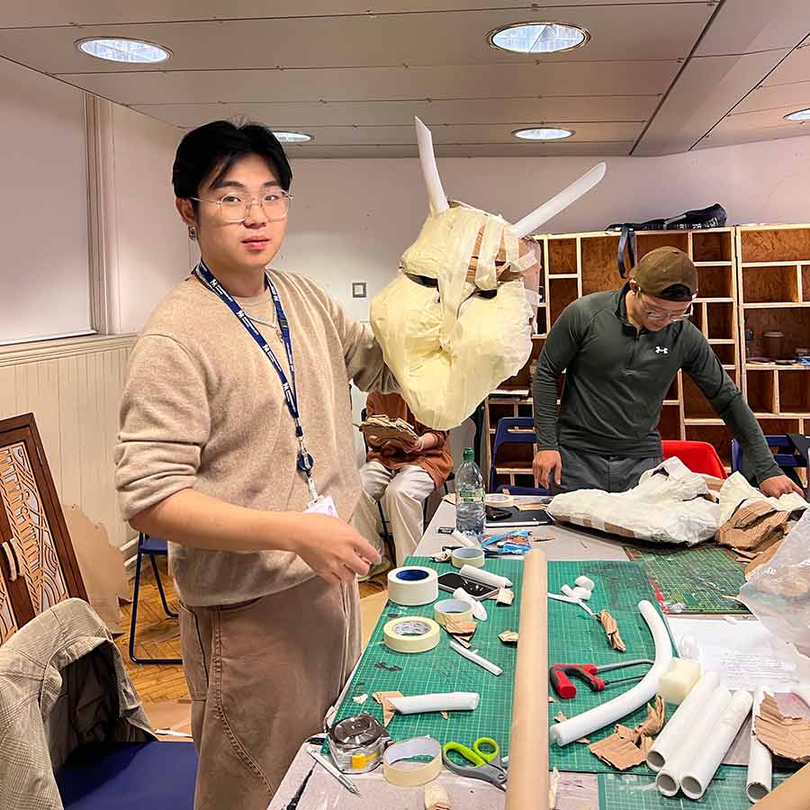 Student holding up sculpture of mask with horns, stood by table with design tools on