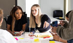 A group of students sits around a table, each holding a phone. Papers and markers are scattered on the table, indicating a collaborative project.