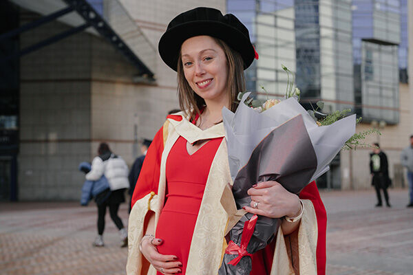 PhD Graduate in a red gown holding her pregnant tummy and flowers