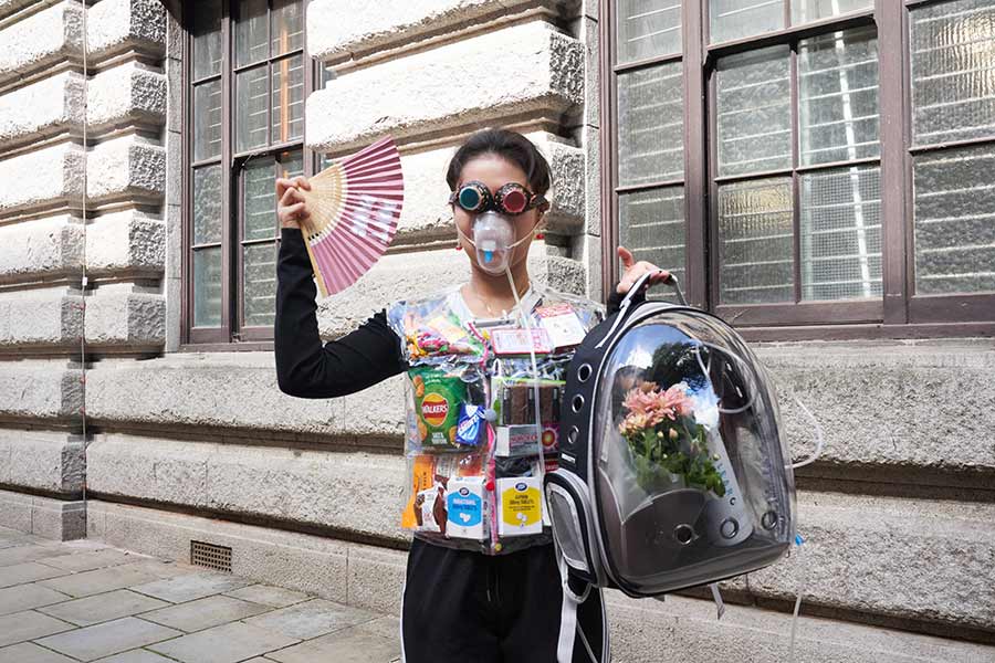 Person stood wearing an oxygen mask, holding a backpack with flowers and oxygen tank in. They are wearing a vest filled with various packaging, including tablets and crisps. They are holding a pink fan and wearing steampunk-like glasses 