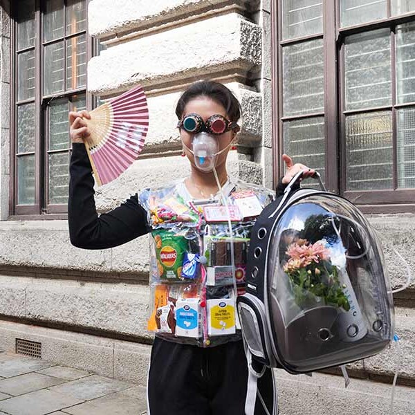 Person stood wearing an oxygen mask, holding a backpack with flowers and oxygen tank in. They are wearing a vest filled with various packaging, including tablets and crisps. They are holding a pink fan and wearing steampunk-like glasses