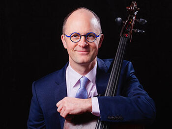 A smiling man with glasses in a blue suit holds a cello.