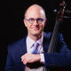 A smiling man with glasses in a blue suit holds a cello.