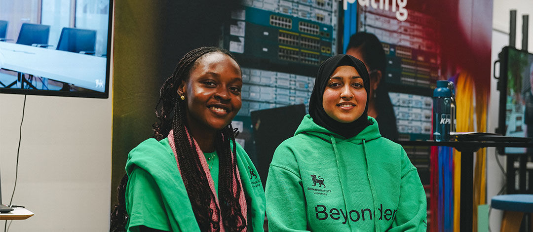 Two female students wearing bright green hoodies which have Beyonder written on them smile at the camera. One student is black with long braided hair pulled back, and the other is wearing a hijab. They're sitting in front of a sign that says computing, suggesting they're student ambassadors at an open day to represent computing courses