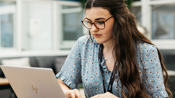 Girl sitting alone at a table working on laptop wearing a 'staff' lanyard