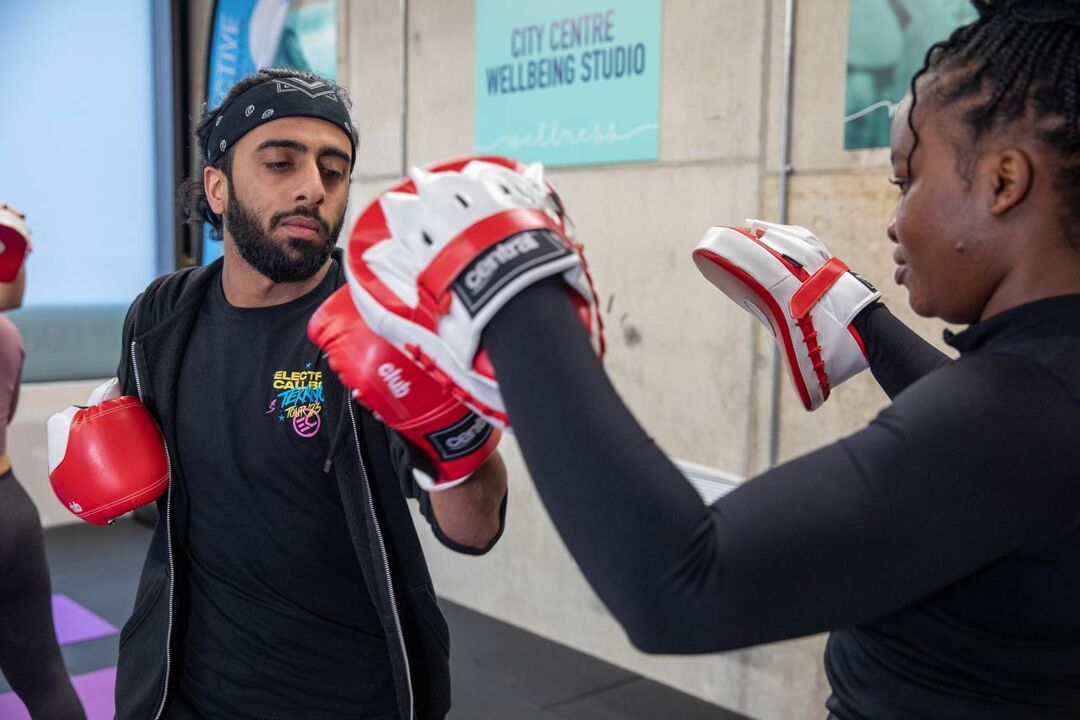 Two students boxing with gloves and pads. A person who appears to be male with light brown skin and a beard faces the camera wearing boxing gloves and punching. A person who appears to be female with dark brown skin and braids faces away from the camera wearing the pads. They are both focused and look relaxed.