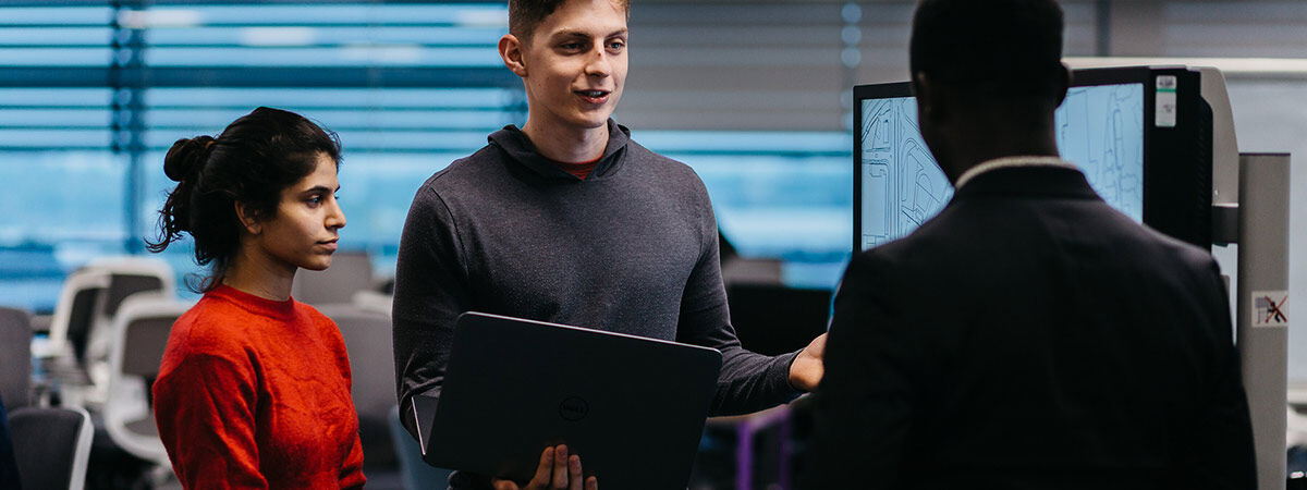 A young man in a grey hoodie holds a laptop and talks to another person while a young woman in a red sweater looks on. A monitor displaying a map is visible in the background.