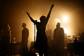 Image from Piaf. Figures in silhouette with female figure in foreground standing at microphone with arms raised.