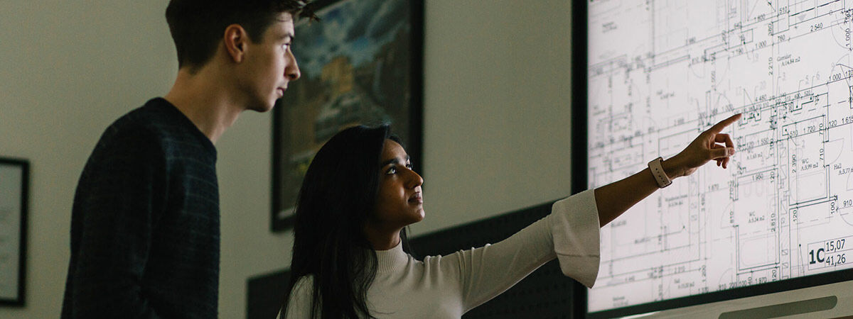 A woman points to a specific section of a large digital architectural blueprint displayed on a screen while a man looks on.