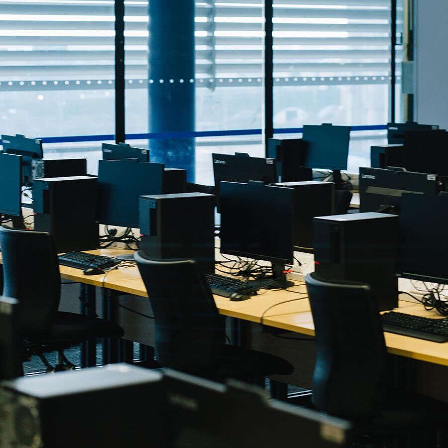 Empty computer lab with multiple workstations, featuring monitors, keyboards, and chairs, arranged on long tables.