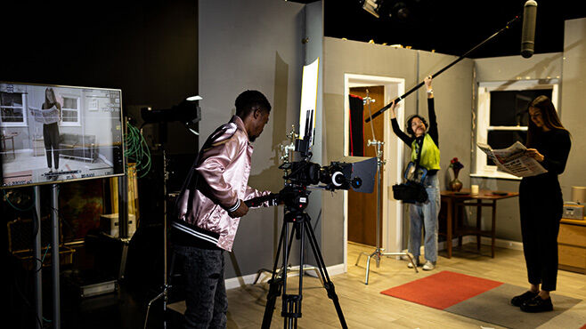 Three students working on video set. One behind camera, one holding mic, one acting