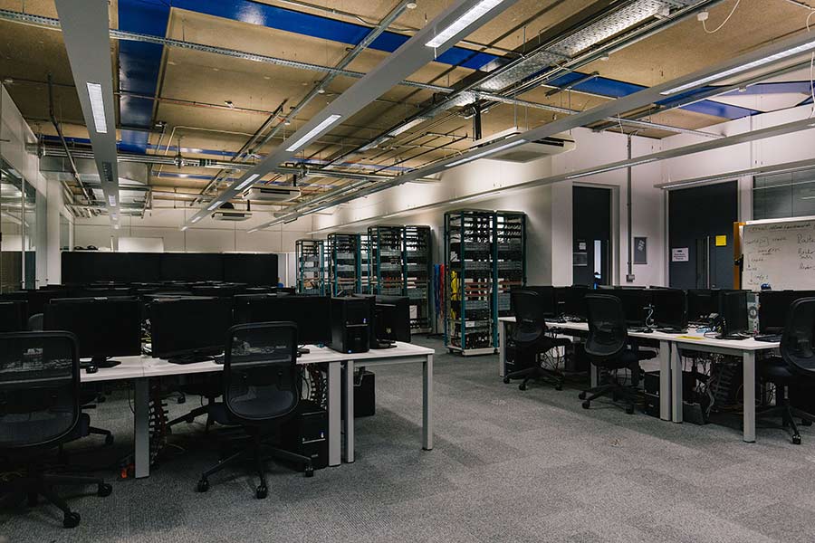 Wide shot of an empty computer lab with rows of workstations, featuring monitors, keyboards, and chairs. Storage racks are visible in the background.