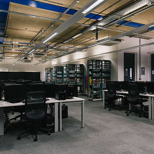 Wide shot of an empty computer lab with rows of workstations, featuring monitors, keyboards, and chairs. Storage racks are visible in the background.