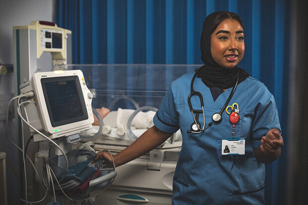 A female healthcare professional wearing blue scrubs, a black hijab, and a stethoscope, stands next to a baby doll in an incubator, with medical equipment on her left.