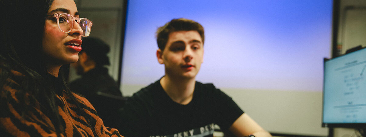 A female student with glasses speaks while a male student in a black t-shirt listens in a computer lab.