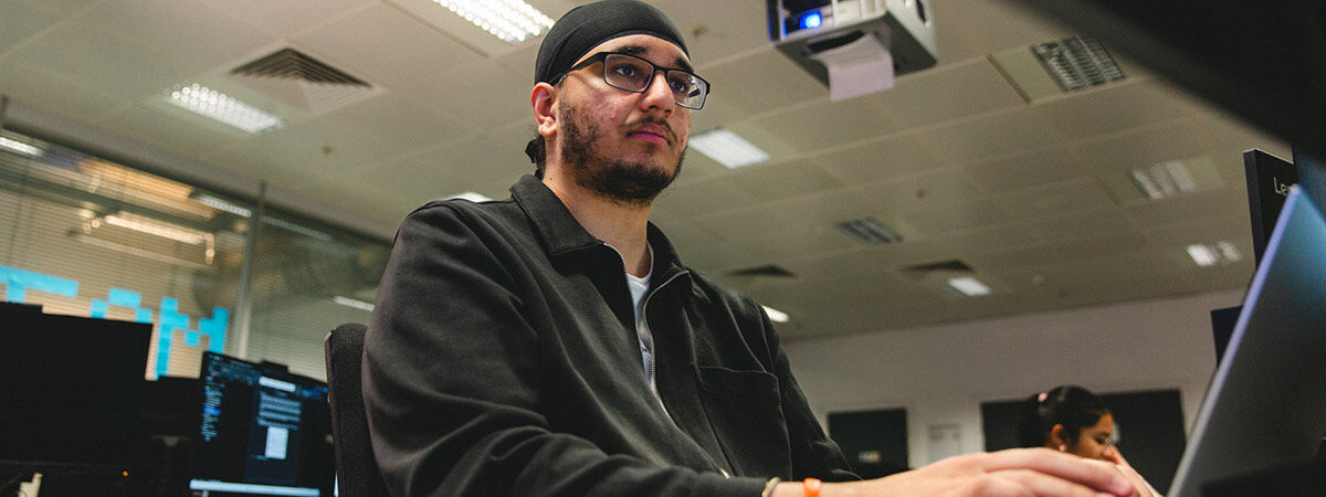 A man wearing a black head covering and glasses sits at a computer desk, looking thoughtfully toward a monitor in a modern computer lab.