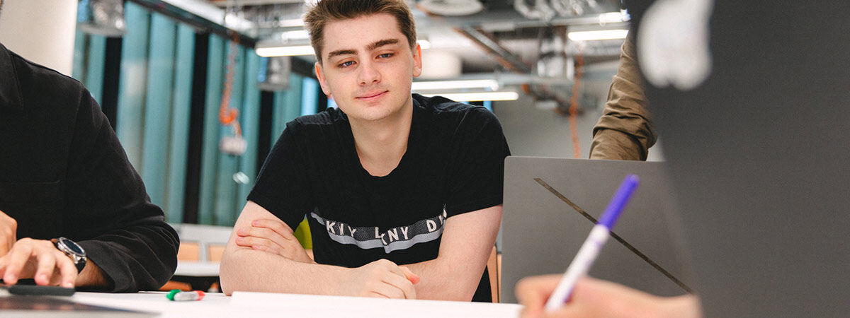 A male student sits at a table with his arms crossed, looking toward a colleague in a bright, industrial-style workspace.