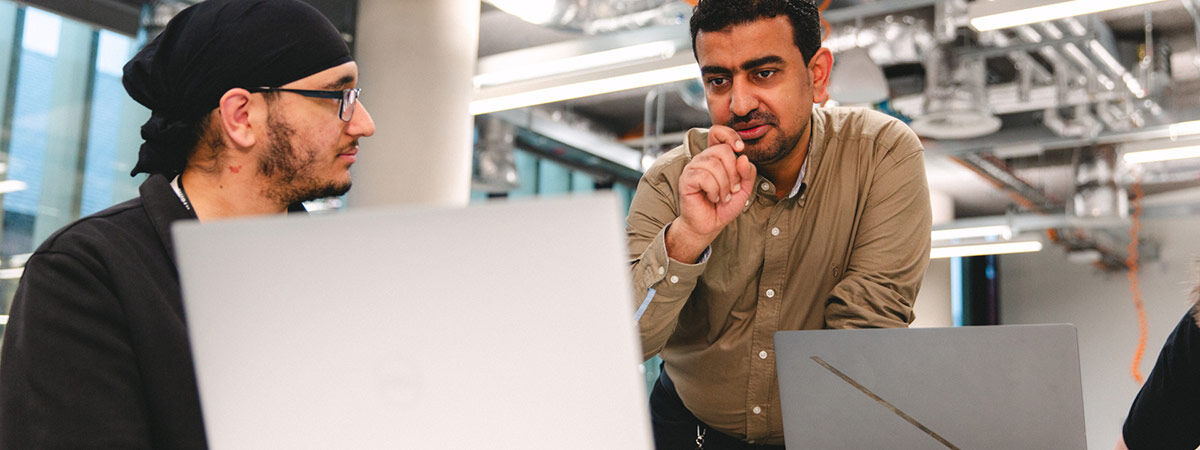 A lecturer gestures while explaining a concept to a student seated at a laptop in a modern office.