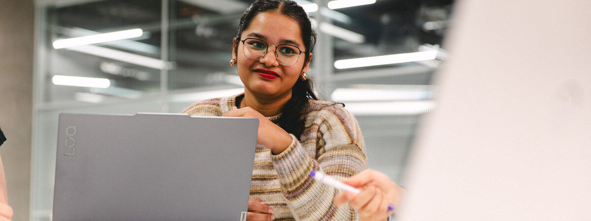A woman wearing glasses and a striped sweater looks toward a colleague while sitting behind a laptop in a brightly lit office.