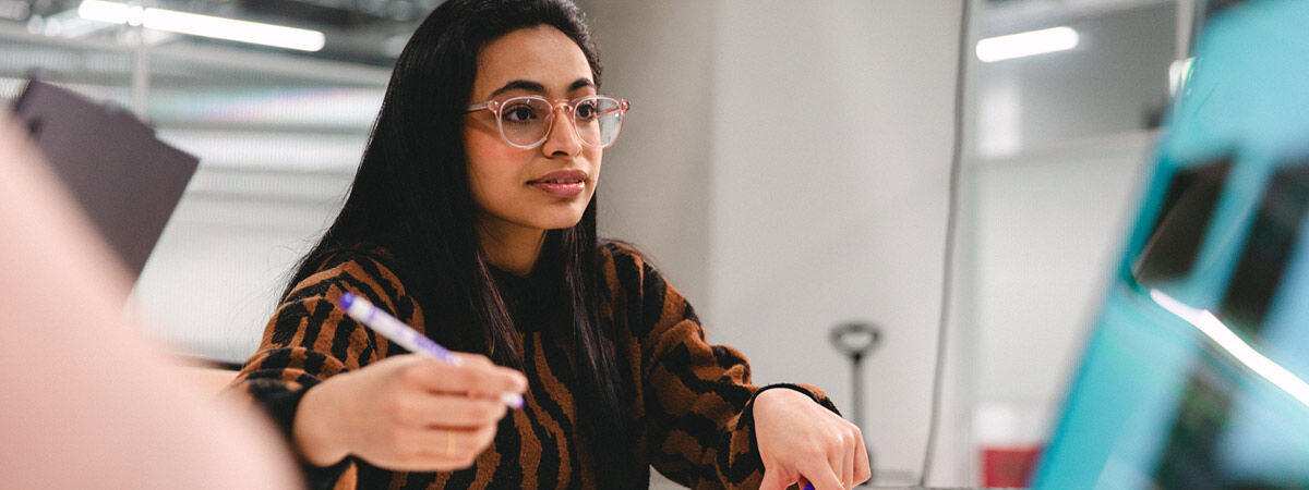 A woman with long dark hair and clear-framed glasses holds a purple marker while looking intently toward a computer screen in a bright, modern lab environment.
