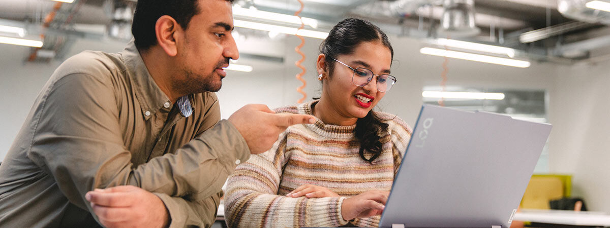 A man points toward a laptop screen while talking to a woman who is smiling and typing in an open office or lab setting.