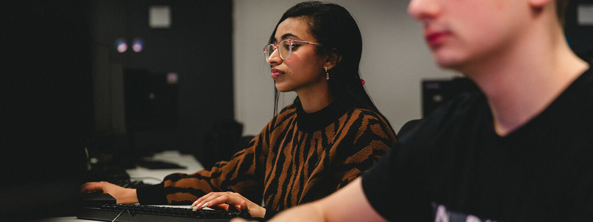A person wearing a tiger-print sweater and glasses works at a computer in a lab or classroom.