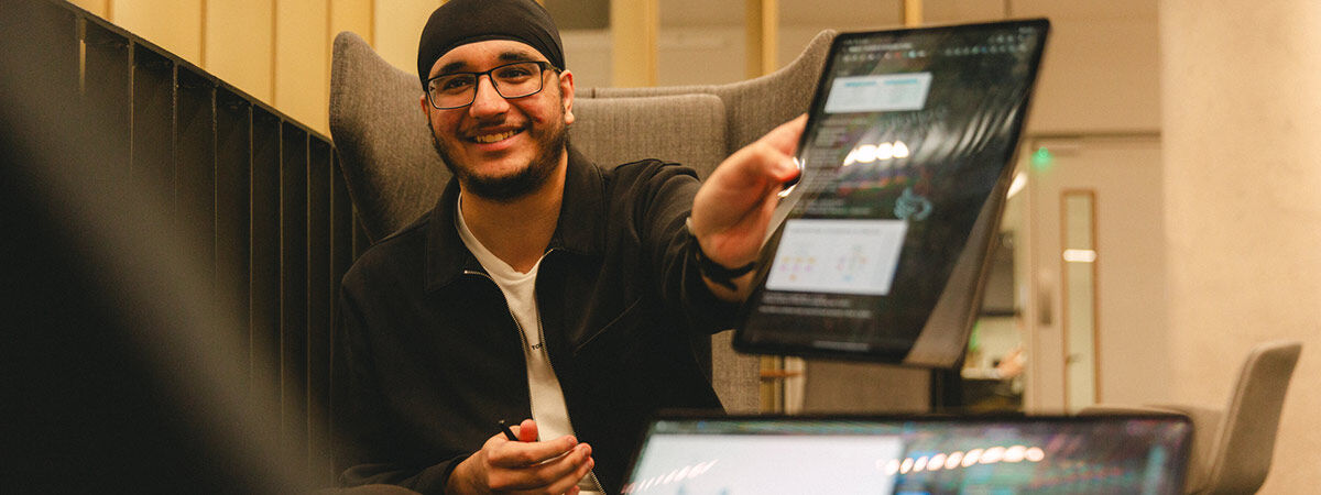 A person wearing glasses and a black jacket smiles while holding up a tablet to show its screen in a modern workspace.