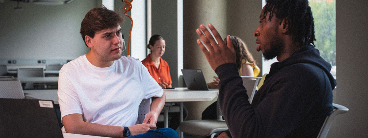 Two male students engage in a discussion at a table in a bright, open-plan workspace while others work in the background.