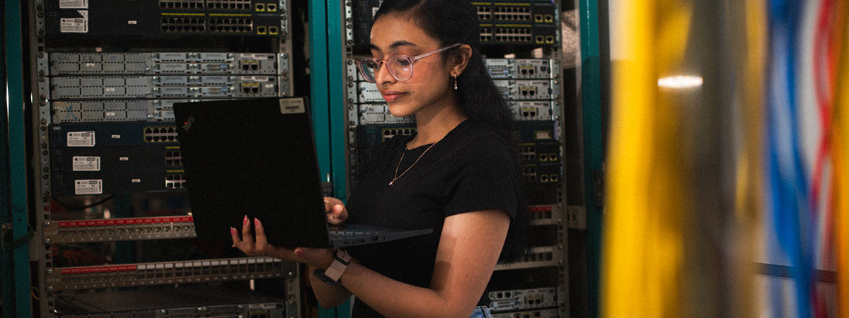 A student wearing glasses holds a laptop while working in front of a rack of network servers.