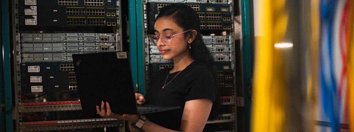 A student is in a server room, holding a laptop