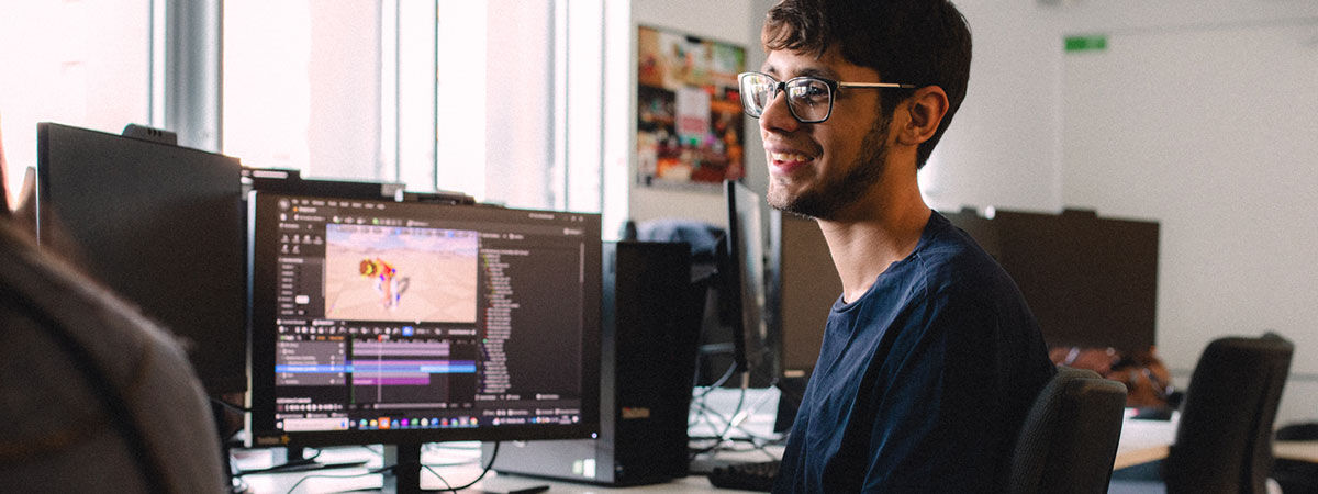 A man with glasses smiles at a computer workstation displaying game development software in a bright, modern lab.