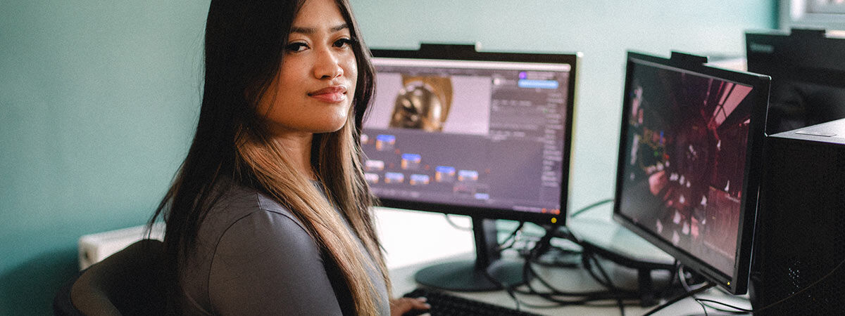  A woman with long dark hair sits at a desk with two computer monitors displaying digital design and game development software.