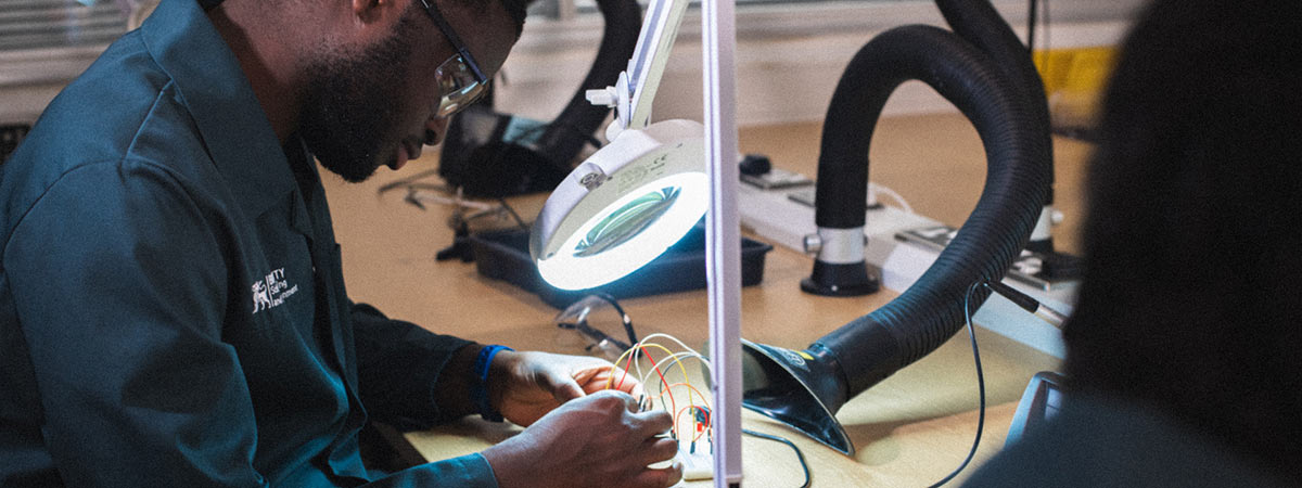 A man in a lab coat and protective eyewear uses a magnifying lamp to meticulously work on an electronic circuit board with various colored wires in a technical laboratory setting.