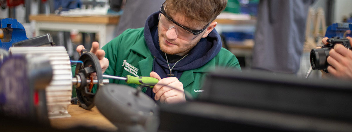 A student in a green lab coat and protective eyewear uses a screwdriver to work on a mechanical component in an engineering workshop.