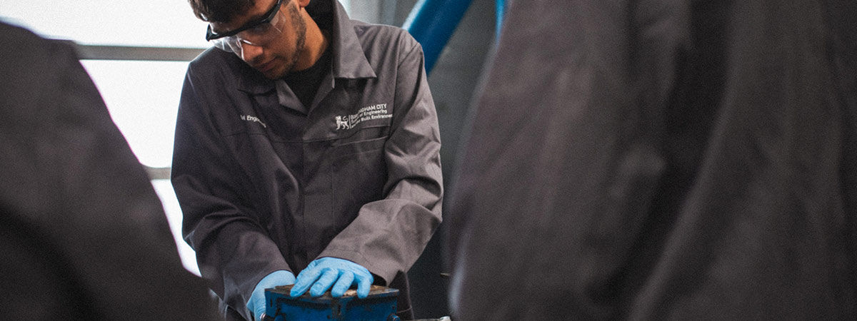 A person wearing protective glasses and a grey lab coat works with blue-gloved hands on a piece of equipment in an engineering workshop.