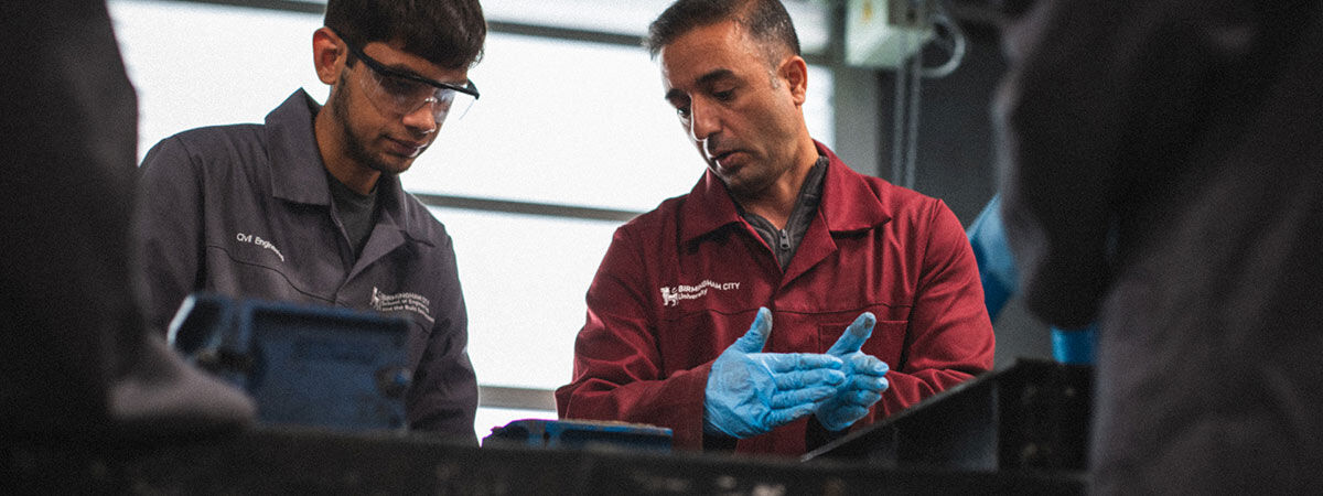 A man in a red lab coat and blue gloves demonstrates a technique to a student in a grey lab coat within an engineering workshop.